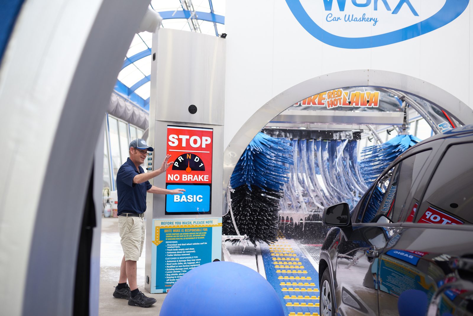 A Brite WorX attendant waving a car onto the conveyor at the tunnel entrance, the wash arch and its brushes running behind him